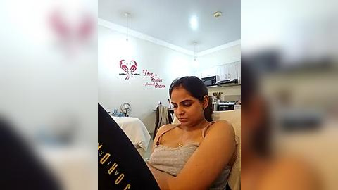 Media: Video of a young woman with medium brown skin, dark hair, and a slim build, wearing a light gray tank top, sitting at a kitchen table in a modern kitchen.