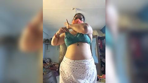 Media: A video of a plus-sized woman in a teal sports bra, pink face mask, and white patterned skirt, adjusting her hair in a cluttered bedroom with clothes and a mirror.