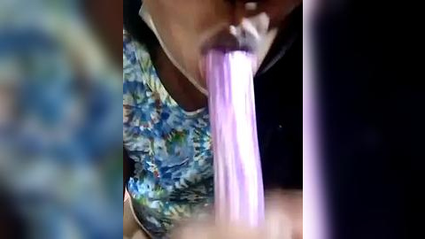 Media: A close-up video of a person with medium brown skin, wearing a floral-patterned blue shirt, using a purple straw to suck on a bubble. The background is blurred, emphasizing the action.