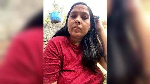 Media: Video of a middle-aged woman with long black hair, wearing a red long-sleeve shirt, looking frustrated, standing outdoors near a blue water container and a stone wall.