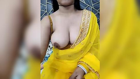 Media: A video of a woman with medium brown skin, wearing a yellow sari with gold embroidery, exposing her large, bare breasts. Her dark hair is partially visible. Background shows a grey geometric pattern.