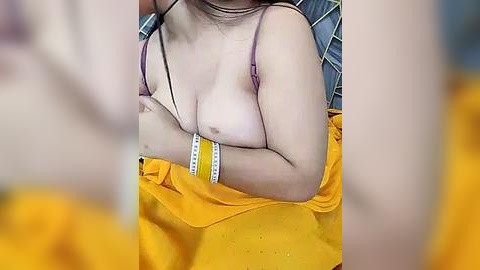 Media: A close-up video of a woman's partially exposed breast, wearing a purple bra and a yellow cloth, with a blurred background.