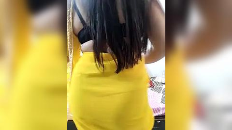 Media: Video of a woman with long, straight black hair, wearing a black bra and a tight yellow skirt, standing in a blurred, brightly-lit room with a bed and patterned sheets in the background.