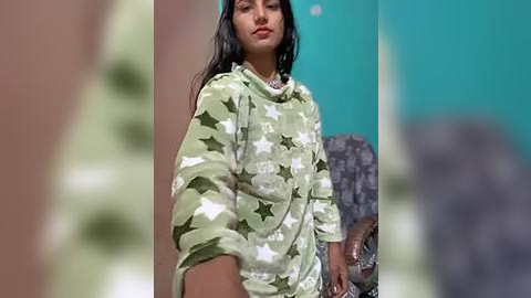 Media: Video of a young South Asian woman with medium brown skin, long black hair, and a confident expression, wearing a green hoodie with white stars. Background features teal and beige walls.