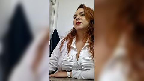 Media: Video of a red-haired woman in a white blouse, looking serious, with a blurred figure in the foreground.