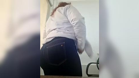 Media: A video of a woman, possibly Latina, standing in a bathroom, wearing a light gray sweatshirt and dark blue jeans, her back to the camera.