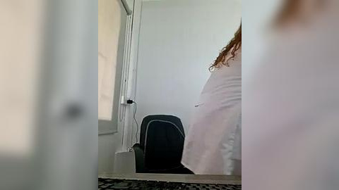 Media: Video of a pregnant woman with curly brown hair, wearing a white dress, standing in a dimly lit, white-walled room with a black chair and a partially open door.