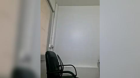 Media: A video of a minimalist room with a black leather chair and white walls, featuring a vertical window with a white curtain and a small, rectangular window on the right. The space appears clean and sparse.