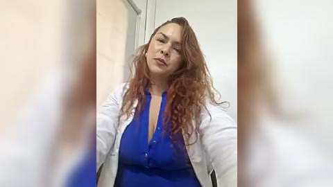 Media: Video of a Caucasian woman with long, wavy red hair, wearing a white lab coat over a blue dress. She has a serious expression. Background includes a blurred white wall and a glass door.