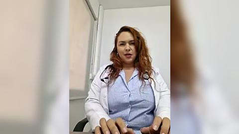 Media: Video of a middle-aged Caucasian woman with curly red hair, wearing a white lab coat and light blue shirt, sitting in an office with white walls and a window.