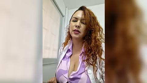 Media: Video of a Caucasian woman with long, curly red hair, wearing a light pink shirt, unbuttoned to reveal cleavage, standing in a sterile room with white walls and a window.