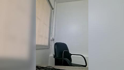 Media: Video of a minimalist office room with a black leather chair against a white wall, a beige window blind, and a white radiator. The scene is dimly lit, creating a calm and sparse atmosphere.