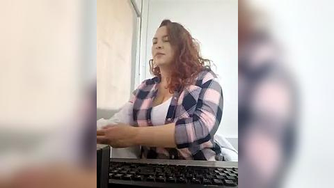 Media: Video of a plus-sized, curly-haired woman in a pink and black plaid shirt, sitting at a desk, typing on a computer, with a blurred figure in the background.