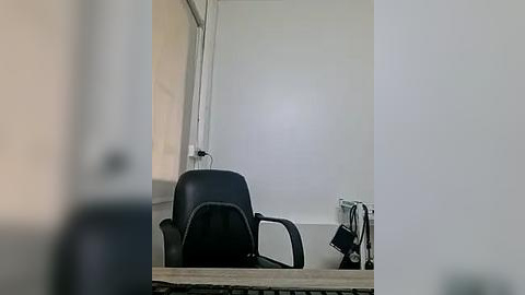 Media: Video of a minimalist office space with a black leather chair, wooden desk, and a white wall with a window. The room is dimly lit, giving a serene, quiet atmosphere.