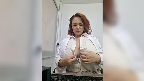 Media: Video of a plus-sized, fair-skinned woman with red hair in a white button-up shirt, unbuttoned to reveal cleavage, seated at a desk in a dimly-lit office.