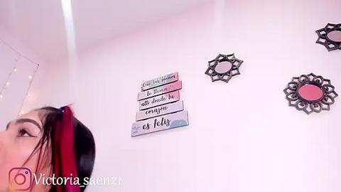 Media: Video of a woman with red and black hair, wearing a black headband, standing in a room with a white wall adorned with decorative letters and a floral wall hanging.