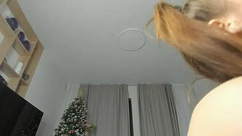 Media: Video of a blonde woman in a white top, seen from behind, in a minimalist living room with a Christmas tree and grey curtains.