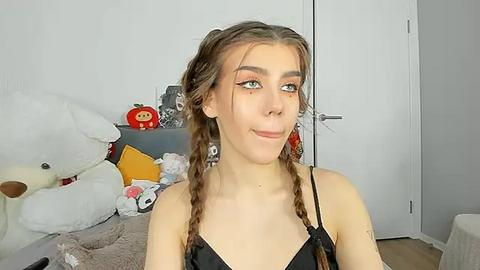 Media: Video of a young Caucasian woman with light skin, brown hair in braids, wearing a black bralette, standing in a cluttered bedroom with stuffed animals and toys.