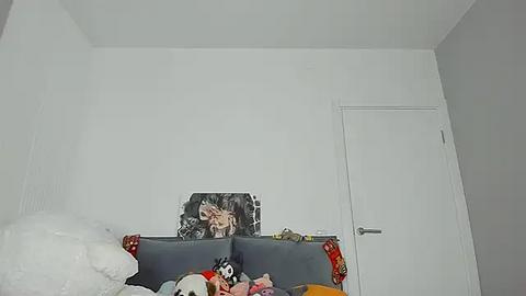 Media: Video of a cluttered bedroom with a white wall, a closed white door, and a bed covered in colorful, messy blankets and pillows. A small, cartoonish figure stands atop the bed.