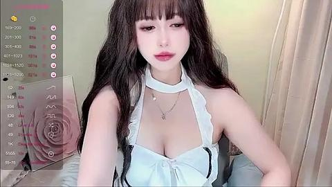 Media: A video of a young Asian woman with long, dark hair and fair skin, wearing a white halter top that accentuates her ample cleavage, in a softly lit, neutral-toned bedroom setting.