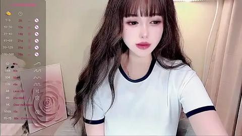 Media: A video of a young Asian woman with long, straight brown hair, wearing a white T-shirt with black trim, sitting in a room with beige curtains and a pink rose on the left.