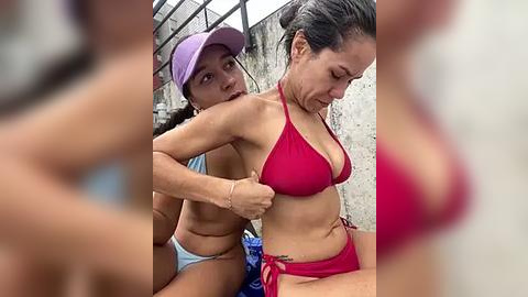 Media: Video of two women in bikinis, one adjusting the other's top strap; outdoor setting with a partially visible structure in the background.