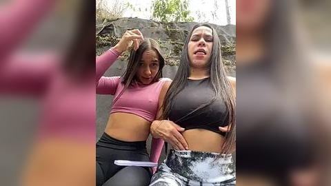 Media: Video of two young women, one with long black hair, in pink crop tops and black leggings, sitting on a grey bench; blurred background.