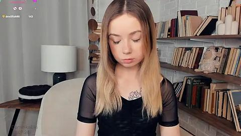 Media: Video of a young, fair-skinned girl with long, ombre blonde hair, wearing a sheer black top and looking down. Background features a white brick wall with bookshelves, a white chair, and a lamp.