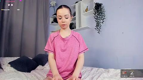 Media: Video of a young Asian girl with light skin, wearing a pink t-shirt, sitting on a bed with floral sheets in a pastel room.