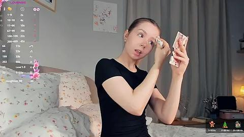 Media: Video of a fair-skinned woman with brown hair in a ponytail, wearing a black shirt, applying makeup in a cozy, dimly-lit bedroom with floral-patterned bedding and curtains.