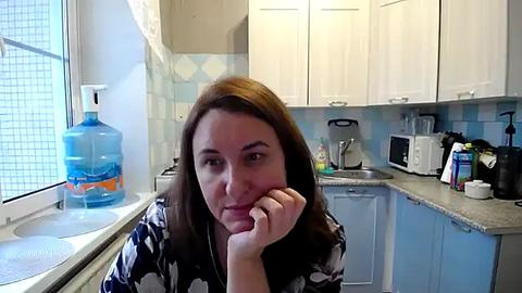 Media: A video of a Caucasian woman with long brown hair, wearing a dark floral robe, leaning on a kitchen counter with a thoughtful expression. The kitchen features white cabinets, a sink, and a water dispenser on the counter.