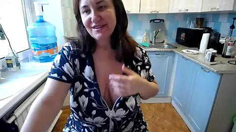 Media: Video of a smiling Caucasian woman with shoulder-length brown hair, wearing a low-cut blue floral dress, exposing cleavage, in a modern kitchen with white cabinets, blue backsplash, and a water dispenser.