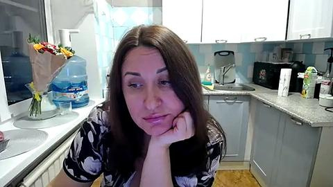 Media: A video of a young woman with long brown hair, wearing a black floral robe, leaning on her elbow in a modern kitchen with white cabinets, a microwave, and a vase of flowers.