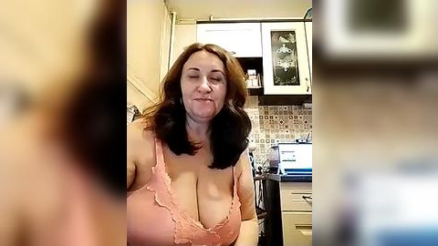 Media: Video of a middle-aged woman with shoulder-length brown hair, wearing a pink lace bra, in a domestic kitchen with white cabinets, a tiled backsplash, and a microwave.