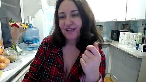 Media: A candid video of a smiling, light-skinned woman with shoulder-length brown hair, wearing a red plaid shirt, standing in a modern kitchen with white cabinets and a granite countertop.