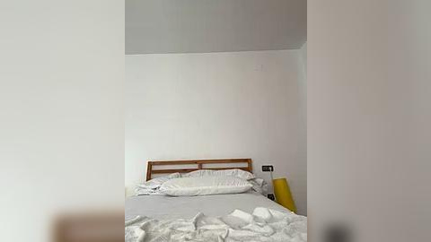 Media: A minimalist bedroom with a white wall, wooden headboard, white bed linens, a yellow pillow, and a partially made bed.