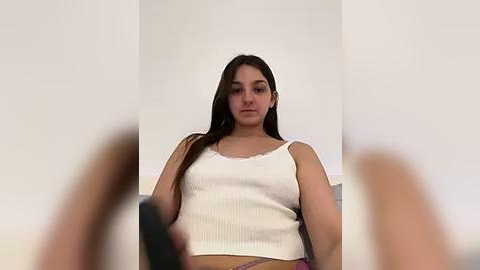 Media: Video of a young Latina woman with long dark hair, wearing a white ribbed tank top, sitting on a gray couch against a plain white wall.