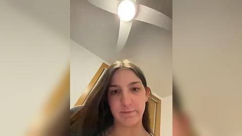 Media: Video of a young woman with long, straight brown hair, light skin, and a neutral expression. She stands in a modern bedroom with a white ceiling fan, wooden door, and beige walls.