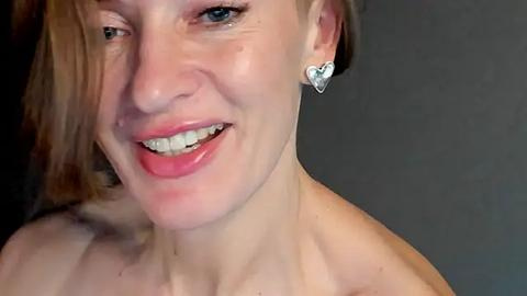 Media: Close-up video of a smiling woman with light skin, short brown hair, and a heart-shaped silver earring, wearing no top, against a plain dark background.