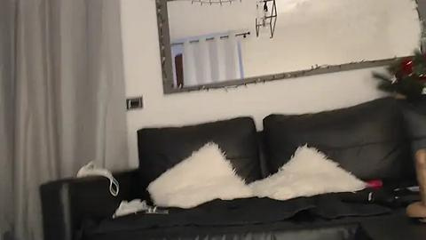 Media: A video of a cozy living room with a black leather sofa, white fur pillows, and a Christmas tree. A mirror with a silver frame hangs on the white wall, and a window with white curtains is visible.