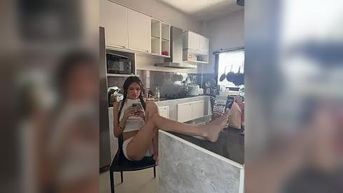 Media: Video of a young woman with long brown hair, wearing a white crop top and panties, sitting on a chair in a modern kitchen, holding a smartphone, legs crossed.