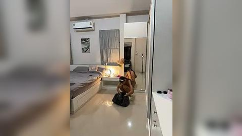 Media: Video of a modern, minimalist bedroom with a beige bed, grey curtains, and a small desk lamp. A large brown bag is on the floor, and an air conditioner is mounted on the wall.