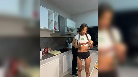 Media: Video of a woman in a kitchen, wearing a white crop top and black shorts, with long hair in braids, standing beside a man wearing black clothes.