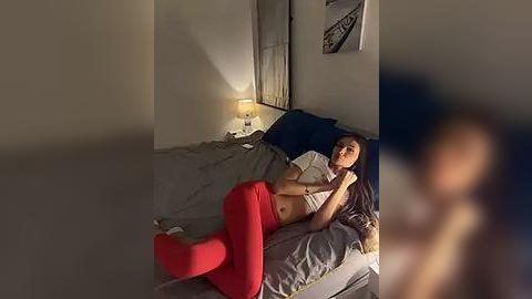 Media: Video of a woman in a dimly lit bedroom, lying on a bed with gray sheets and red pants.