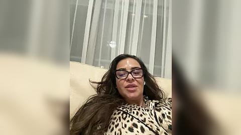 Media: Video of a woman with long brown hair, wearing glasses and a leopard-print coat, lying on a beige sofa, surrounded by sheer white curtains in a modern living room.