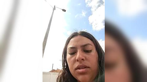 Media: A low-angle video of a young woman with wet, dark hair, focused eyes, and a neutral expression, under a partly cloudy sky with a tall wind turbine in the background.