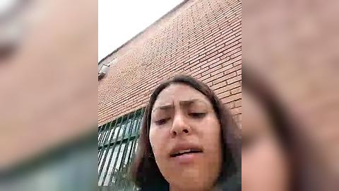 Media: A video of a woman with brown skin and straight black hair, looking distressed, standing in front of a red-brick building with a green window.