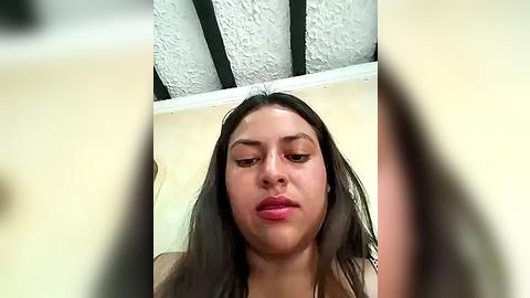 Media: Video of a young woman with long dark hair, pale skin, and full lips, wearing minimal makeup, against a textured white ceiling and beige wall.