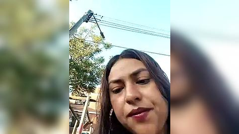 Media: Video of a young woman with medium-length brown hair, wearing minimal makeup, looking downward with a neutral expression. The background features a blurry urban scene with power lines, trees, and a wooden fence.