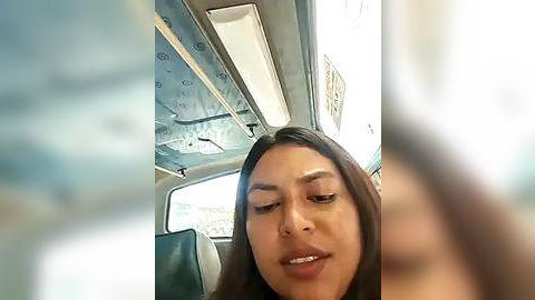 Media: Video of a Latina woman with long brown hair, slightly smiling, seated inside a dimly lit, old-fashioned bus. The interior is worn, with visible ceiling patterns and a window view of an outdoor scene.